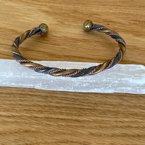 May include: A twisted metal cuff bracelet with bronze, copper, and black tones. The bracelet has a braided design and rounded end caps. The jewelry is displayed on a white rectangular object, set against a wooden background.