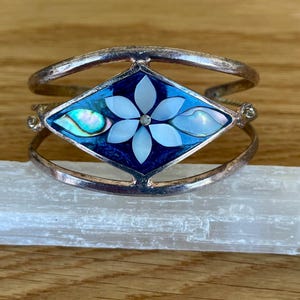 May include: Silver-toned cuff bracelet with a central diamond-shaped design. The design features a blue background with a white flower and iridescent accents. The bracelet is open and rests on a white crystal.