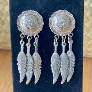 May include: Silver-toned earrings with a circular top featuring a sunburst design and a scalloped edge. Three dangling feather charms hang from each earring. The earrings are displayed against a black background.