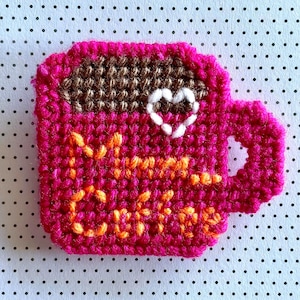 May include: A handmade, crocheted coffee mug in shades of pink, brown, and orange. The mug features a white heart and the words "More Coffee" stitched on the front. The mug has a handle and is set against a dotted background.