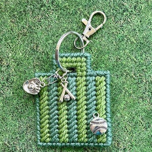 May include: A green and blue stitched keychain with baseball-themed charms. The keychain features a baseball glove, baseball bat and ball, and a silver clasp for easy attachment. The background is green.