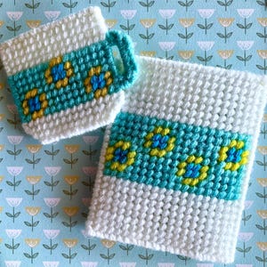 May include: A white crocheted mug and coaster set. The mug has a turquoise handle and a turquoise band with yellow and blue floral designs. The coaster has a matching turquoise band with the same floral pattern. The set is on a light blue floral background.