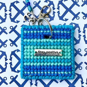 May include: A square, hand-stitched keychain in shades of blue and turquoise, with the words "Stay positive" on a silver plate. The keychain has a silver clasp and an anchor charm, with a blue beaded lanyard.