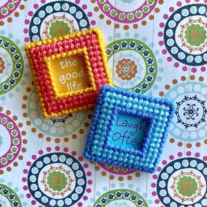 May include: Two handmade, square needlepoint pieces. One is red and yellow with the words "the good life." The other is blue with the words "Laugh Often." Both are on a patterned background.