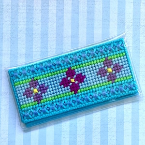 May include: A turquoise and blue needlepoint checkbook cover with a floral design. The cover features three purple flowers with yellow centers, set against green and blue stripes. The cover is encased in a clear plastic sleeve.
