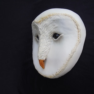 May include: A white owl mask with a detailed, textured surface. The mask features dark eyes, a tan beak, and a decorative trim around the edges. The owl mask is set against a black background.