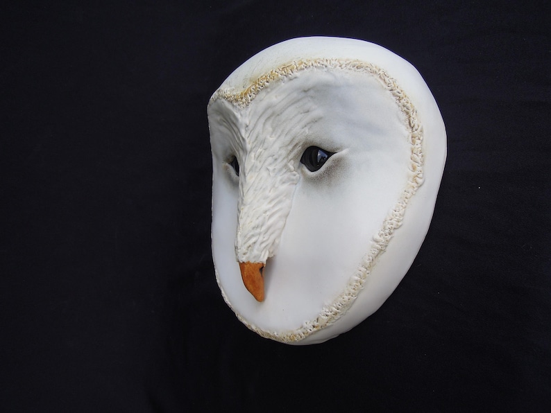 May include: A white owl mask with a detailed, textured surface. The mask features dark eyes, a tan beak, and a decorative trim around the edges. The owl mask is set against a black background.