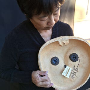 May include: A beige ceramic bowl with a face design, featuring two dark blue eye-like shapes and a small, attached tag with handwritten text. The bowl is held by a person wearing a black sweater.