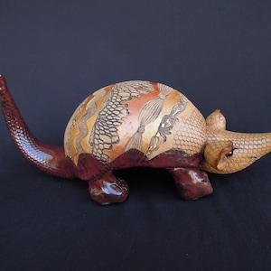 May include: A decorative armadillo figurine crafted from a gourd, featuring a glossy dark reddish-brown tail and legs. The shell displays intricate pyrography patterns in tan and orange, including geometric scales, wavy lines, and floral motifs.