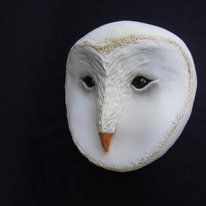 May include: A white owl mask with a detailed, textured surface. The mask features dark eyes, a tan beak, and a textured border. The owl's face is centered against a black background, creating a striking contrast.