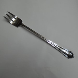 May include: A silver fork with three tines, angled slightly to the left. The handle has a decorative design near the end. The fork is made of a shiny, reflective metal, likely stainless steel. This utensil is suitable for serving appetizers or desserts.