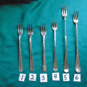 May include: A set of six silver-toned metal forks arranged in a row, numbered one through six on small white cards. The forks vary in length and handle design, with some featuring plain stems and others displaying intricate embossed patterns.