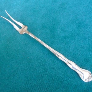 May include: A silver-toned metal cocktail fork featuring two long, slender tines and a smooth, elongated handle that terminates in a decorative, rounded end with subtle embossed scrollwork details along the neck and base of the handle.