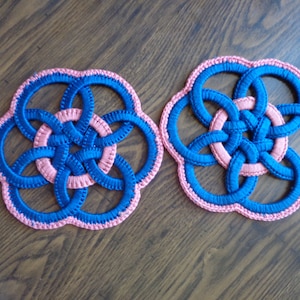 May include: Two crocheted coasters in a blue and pink color scheme. The coasters are circular and feature a woven design with multiple loops.