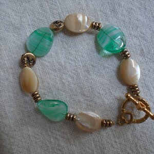 May include: A bracelet with green and white oval beads, gold toned spacers, and a gold toned toggle clasp.