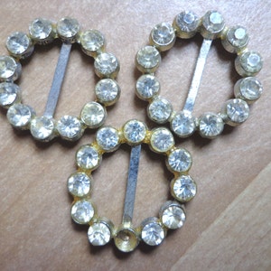 Puede incluir: Tres hebillas de metal plateadas con adornos de diamantes de imitación transparentes. Cada hebilla tiene una forma redonda y una barra de metal que se desliza por el centro.