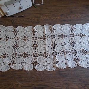 May include: Cream-colored, rectangular crocheted doily with a repeating floral pattern. The openwork design features a series of connected flower-like shapes, creating a delicate and intricate texture. The doily is displayed on a dark wooden surface.