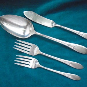 May include: A set of four silver-toned metal utensils including one large oval spoon, one butter knife with a rounded blade, and two forks with four tines each. Each piece features a slender handle with a small, engraved floral motif.