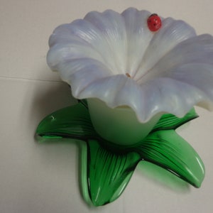 Pode incluir: Uma flor de vidro branco com uma base de vidro verde. Uma pequena joaninha vermelha está pousada na borda da flor.