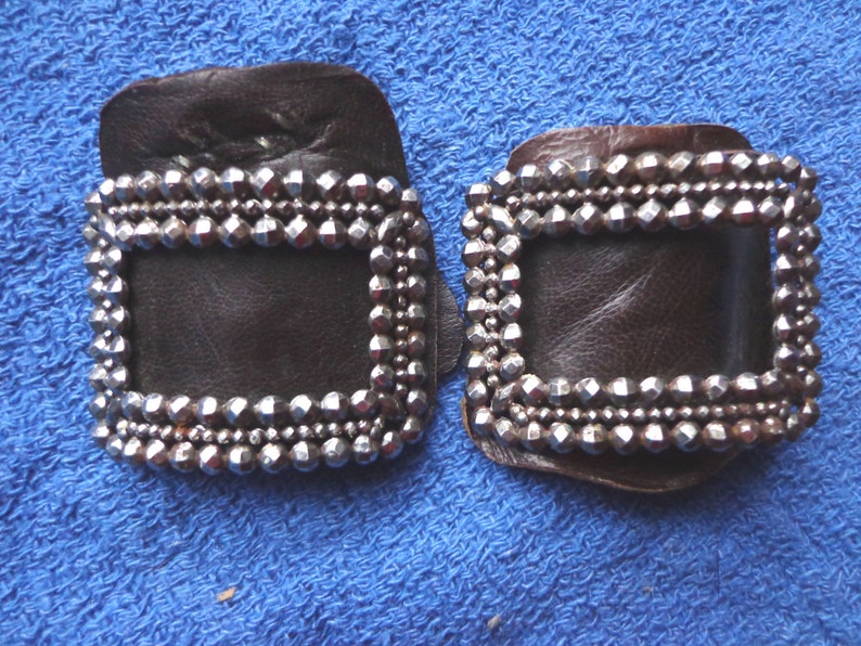 Puede incluir: Dos hebillas de cuero marr&oacute;n con tachuelas de metal plateado. Las hebillas son rectangulares con una abertura cuadrada en el centro.