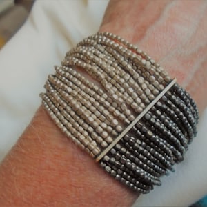 May include: A silver beaded bracelet with a silver bar clasp. The bracelet is made of two shades of silver beads, one light and one dark.