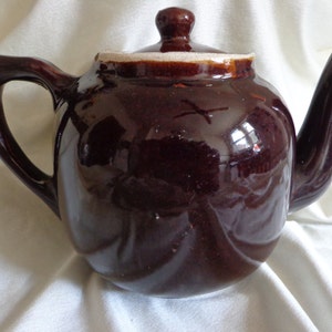 May include: A brown ceramic teapot with a white lid and a white rim around the spout. The teapot has a rounded body and a handle on the left side.