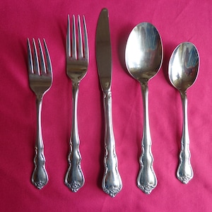 May include: A set of five silver-plated utensils, including a knife, fork, and three spoons, arranged on a pink surface.