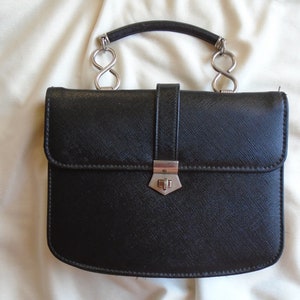 Puede incluir: Un bolso de mano de cuero negro con un cierre de plata y un asa de plata con detalles de símbolo de infinito.