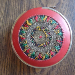 Puede incluir: Una cinta métrica de costura redonda roja con un borde de metal dorado y un diseño circular bordado en hilo dorado, verde y rojo.