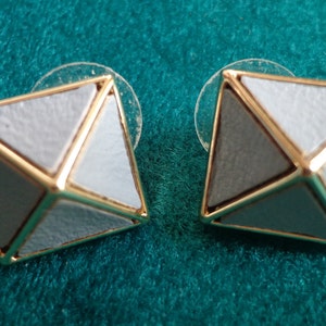 Pode incluir: Um par de brincos dourados com um design geométrico. Cada brinco tem a forma de uma pirâmide com três lados de esmalte branco e um lado de esmalte cinza.
