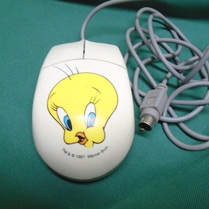 Könnte beinhalten: Eine weiße Computermaus mit einer gelben Tweety Bird-Cartoonfigur auf der Oberseite. Die Maus hat ein graues Kabel und einen PS/2-Stecker. Der Text "TM & © 1997 Warner Bros." ist auf der Unterseite der Maus aufgedruckt.