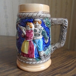 May include: A ceramic beer stein with a tree branch handle. The stein features a raised relief design of a couple in traditional clothing, set against a blue background with white polka dots.