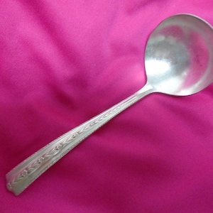 Puede incluir: Un cucharón de plata con un mango adornado. El cucharón está sobre un fondo de tela rosa.