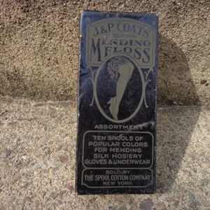 Puede incluir: Una caja negra con letras doradas que dicen "J&P Coats Scented Mending Floss Assortment Ten Spools of Popular Colors for Mending Silk Hosiery Gloves & Underwear Sold by The Spool Cotton Company New York". La caja presenta una ilustración en blanco y negro de una pierna de mujer.