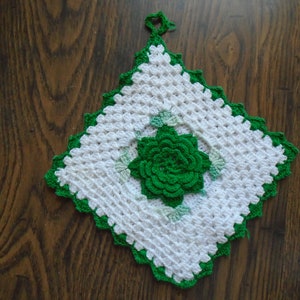 May include: A white crocheted square with a green crocheted flower in the center. The square has a green crocheted border.