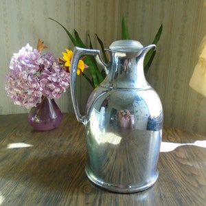 May include: A silver-coloured, vintage-style, thermal carafe with a glass lid. The carafe has a rounded body and a handle. It is sitting on a wooden table.