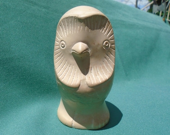 May include: A carved stone owl sculpture. The owl is light beige with etched details, including the eyes, beak, and feather patterns. The sculpture is standing upright on a teal surface.