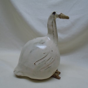 May include: A white ceramic goose figurine with brown accents. The goose has a long neck and a rounded body.