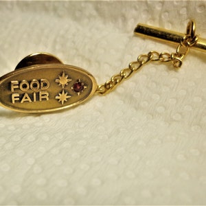 以下が含まれることがあります： 赤い石と2つの星が付いたゴールドトーンのラペルピン。ピンには「FOOD FAIR」と書かれています。