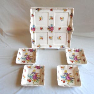 May include: A vintage porcelain serving set featuring a large rectangular tray and four square dishes. The set is decorated with colorful floral patterns in pink, purple, yellow, and blue, set against a cream background.