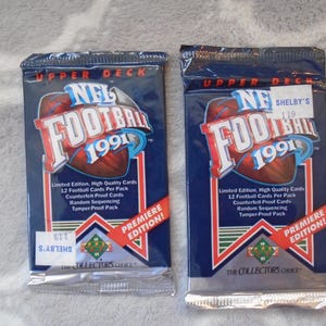 May include: Two sealed packs of 1991 Upper Deck NFL Football cards. The packs are navy blue with a football graphic and the text "NFL FOOTBALL 1991". Each pack is labeled "Premiere Edition!" and "The Collector's Choice".