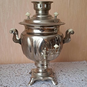 May include: A silver samovar with a decorative design and ivory-colored knobs. The samovar is sitting on a white lace tablecloth.