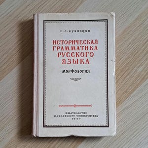 May include: A vintage book titled "Historical Grammar of the Russian Language. Morphology" in Russian. The cover is off-white with red and black text. The book is lying on a wooden surface.