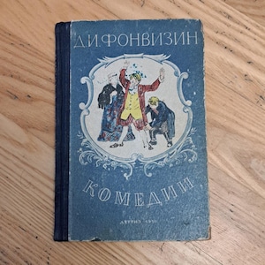 May include: A vintage book with a blue cover featuring the title "Д.И.ФОНВИЗИН" and the word "КОМЕДИИ" in Cyrillic script. The cover illustration depicts three figures in period clothing. The book is set on a wooden surface.