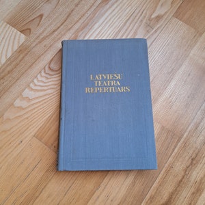 May include: A gray hardcover book with the title "LATVIEŠU TEATRA REPERTUĀRS" in gold lettering.