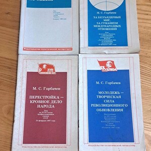 Op de afbeelding: Vier blauwe en rode paperbacks met witte tekst. De boeken zijn getiteld "Sneller herstructureren, op een nieuwe manier handelen", "Voor een nucleaire wereld, voor humanisme in internationale betrekkingen", "Perestroika - de zaak van het volk", en "Jeugd - de creatieve kracht van de revolutionaire vernieuwing".
