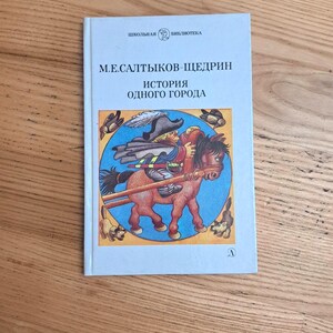 May include: A hardcover book titled "История одного города" (History of a Town) by M.E. Saltykov-Shchedrin. The cover features an illustration of a person riding a horse, with text in Russian. The book is from the "School Library" series.