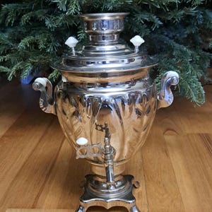May include: A polished silver samovar with a decorative design, featuring a spigot, handles, and a lid with white knobs. The samovar stands on a decorative base. The background includes green pine branches.
