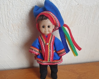 May include: A small doll wearing a blue and red traditional costume with a blue hat and colorful tassels. The doll has brown hair and brown eyes.