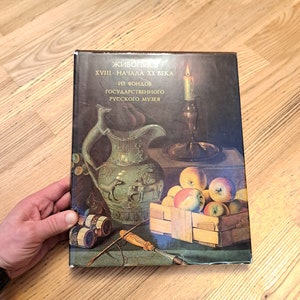 Op de afbeelding: Een boekomslag met een schilderij van een stilleven met een groene keramische kan, een kandelaar met een brandende kaars en een mand met appels. De titel van het boek is "Живопись XVIII - Начала XX века из фондов Государственного Русского музея".